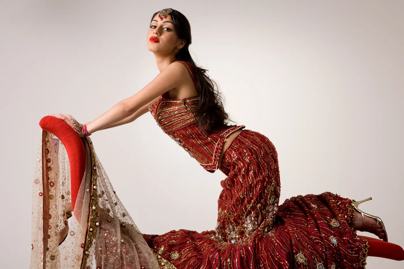 Woman in a red and gold embellished lehenga kneeling on a red velvet chaise, looking over her shoulder toward the camera.