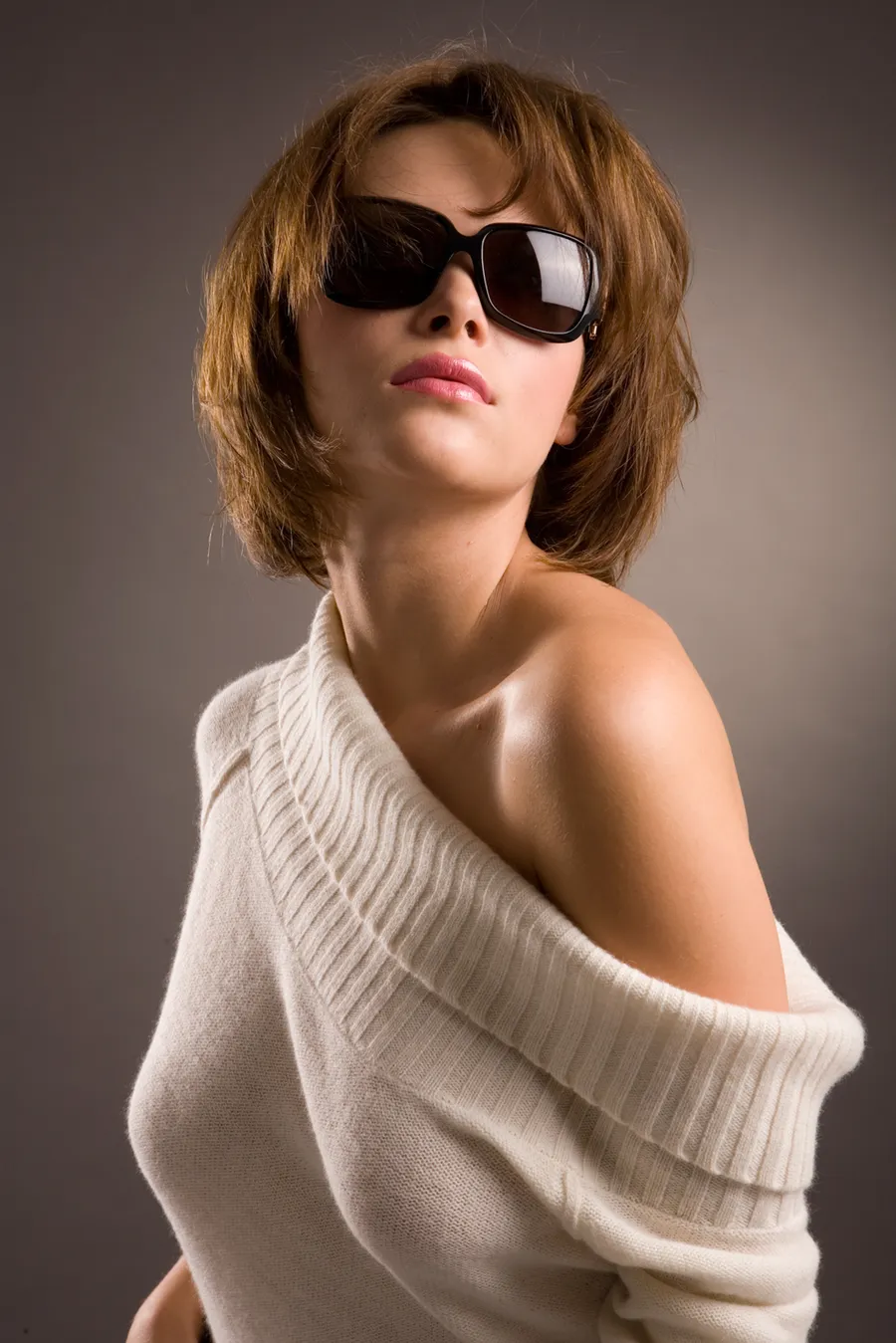 Woman in a black satin-strap top with wraparound sunglasses, arms crossed at her chest in a dark studio portrait.