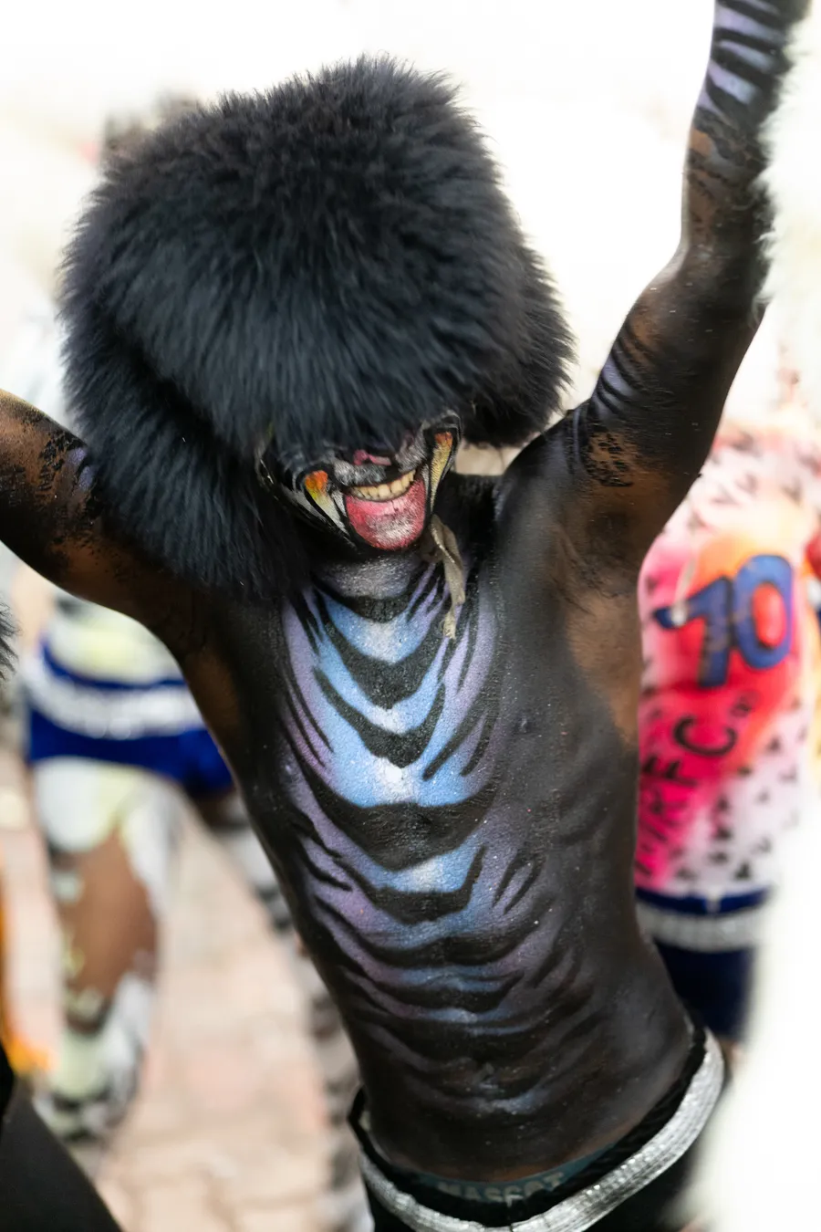 Black-painted Pili Vesha performer with blue tiger-inspired stripes raises both arms beneath a furry black headpiece.