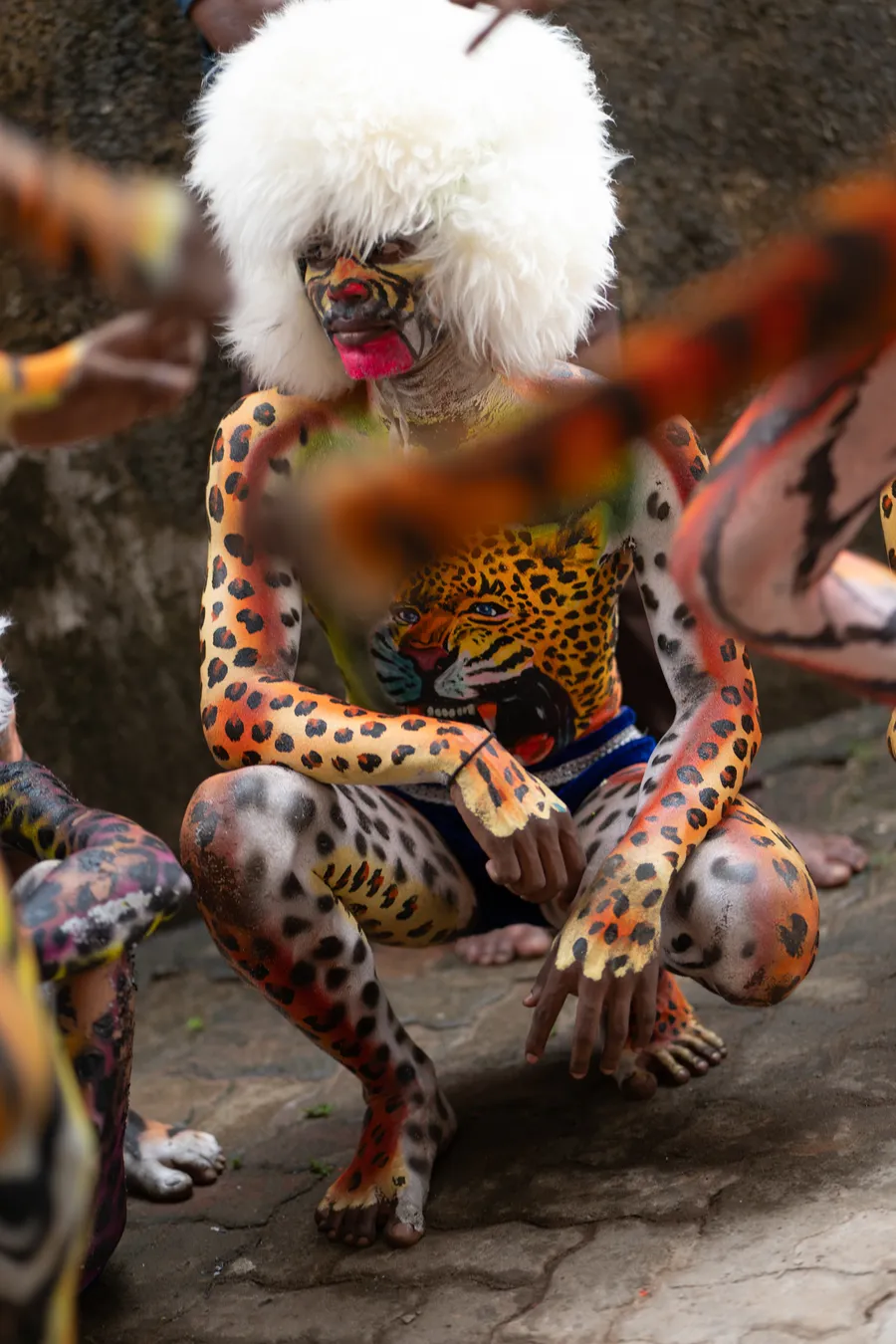 Seated Pili Vesha performer in spotted body painting and a white furry headpiece, framed by other performers.