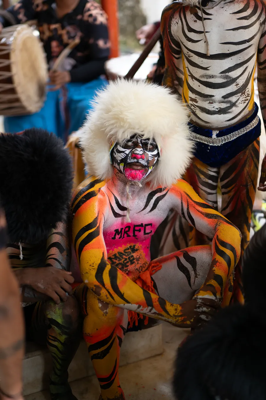 Seated Pili Vesha performer with orange-striped body painting and a white furry headpiece beside a drummer.