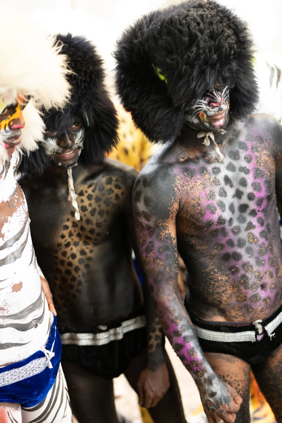 Two black-painted Pili Vesha performers in furry black headpieces, one with spotted purple body painting.