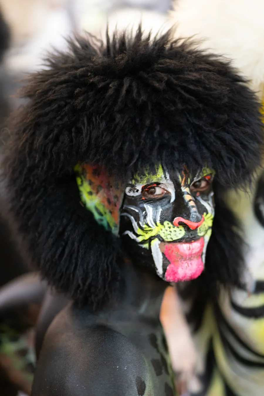 Close view of a performer in a furry black headpiece with green, white, and pink tiger face paint.