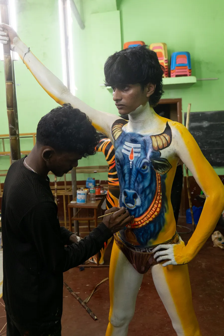 Artist paints a blue animal face on the torso of a yellow-and-white body-painted performer.