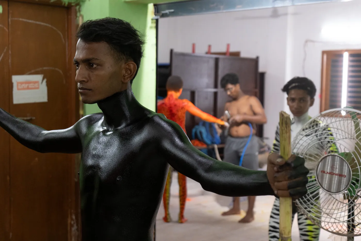 Black-painted performer stretches between bamboo poles while other performers prepare in the room.