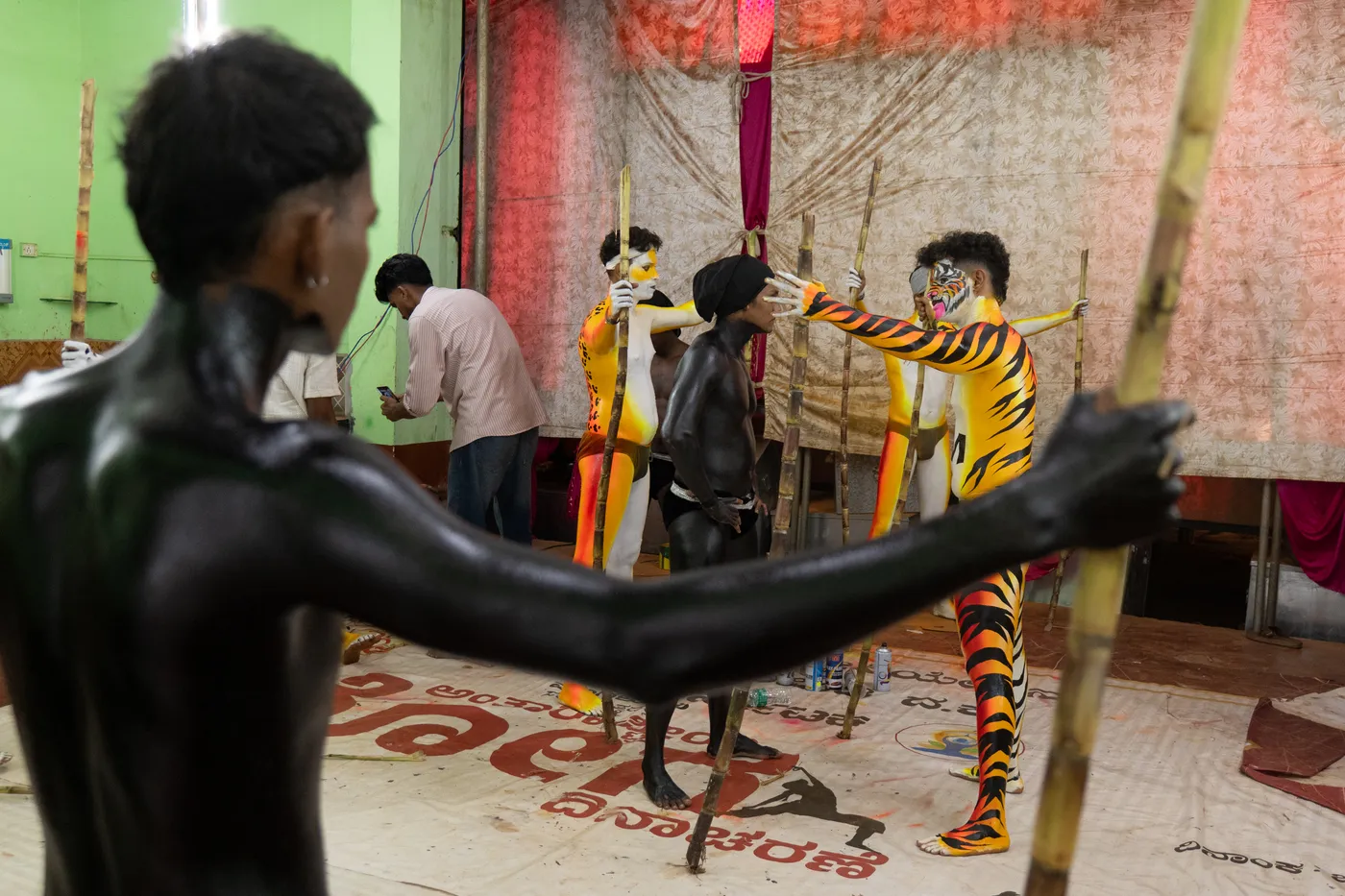 Several body-painted performers hold bamboo poles while posing in a preparation room.