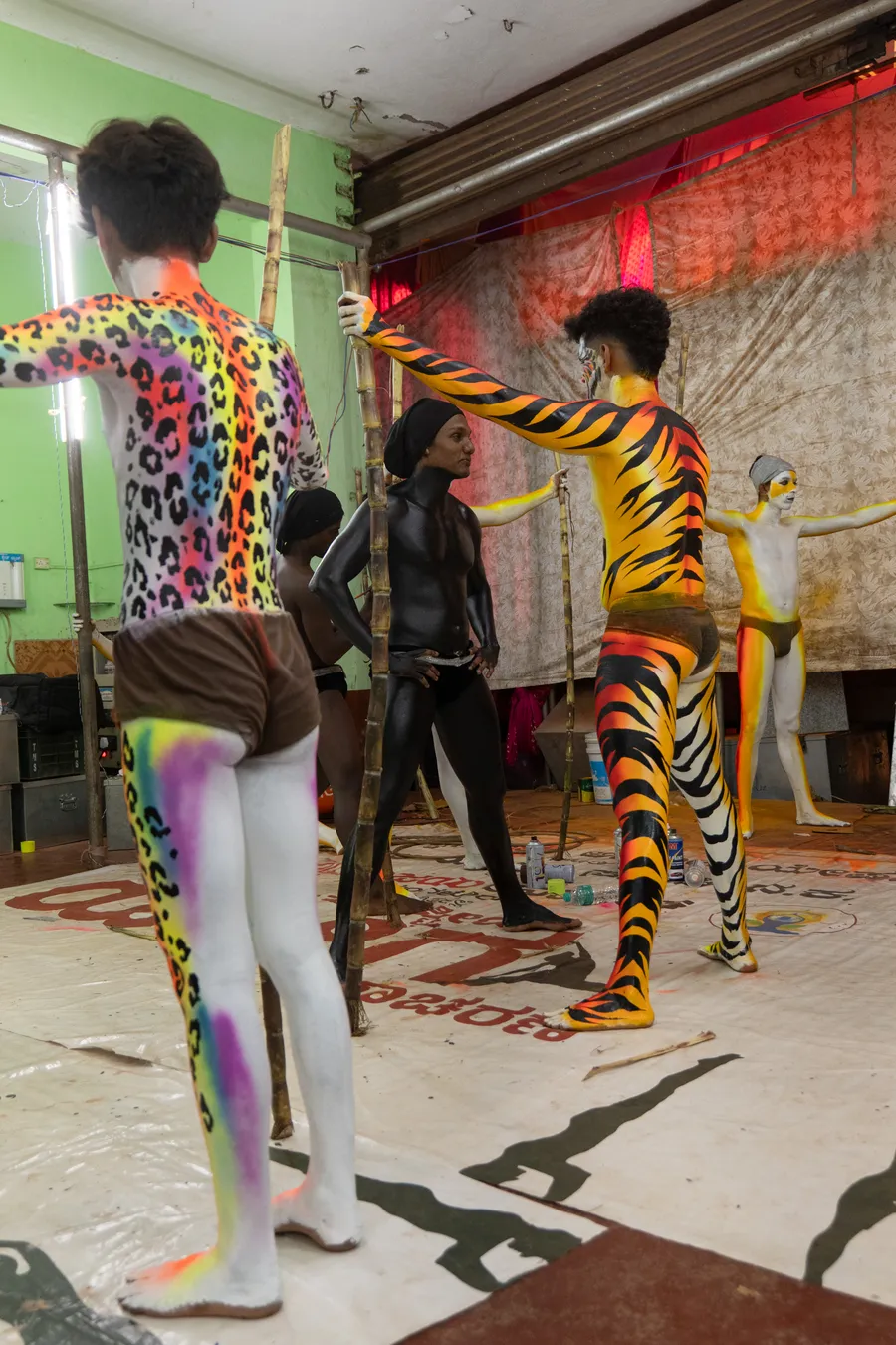 Body-painted performers stand with bamboo poles in a room lined with paint supplies.