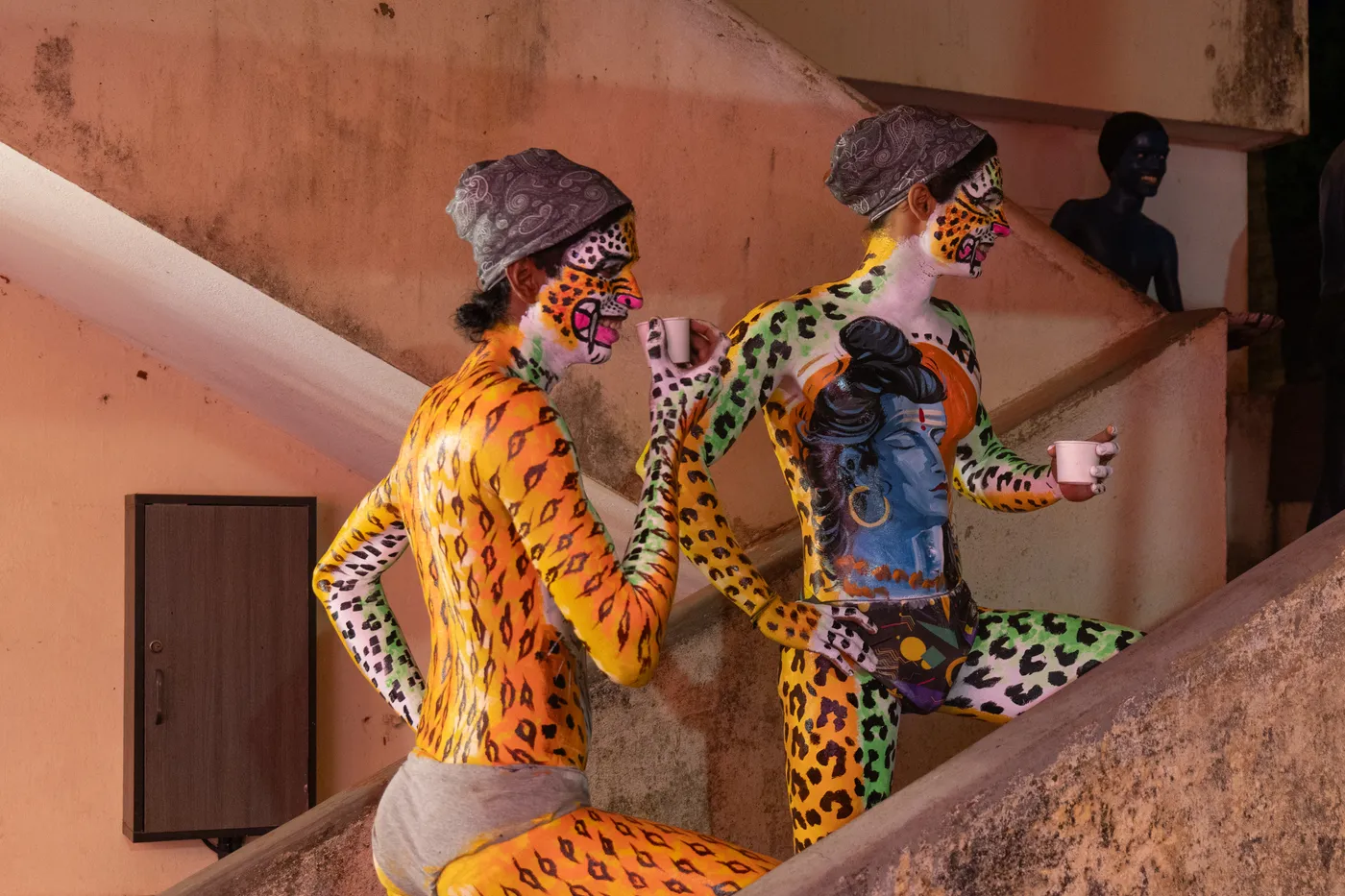 Two spotted body-painted performers hold paper cups on a concrete stairway.