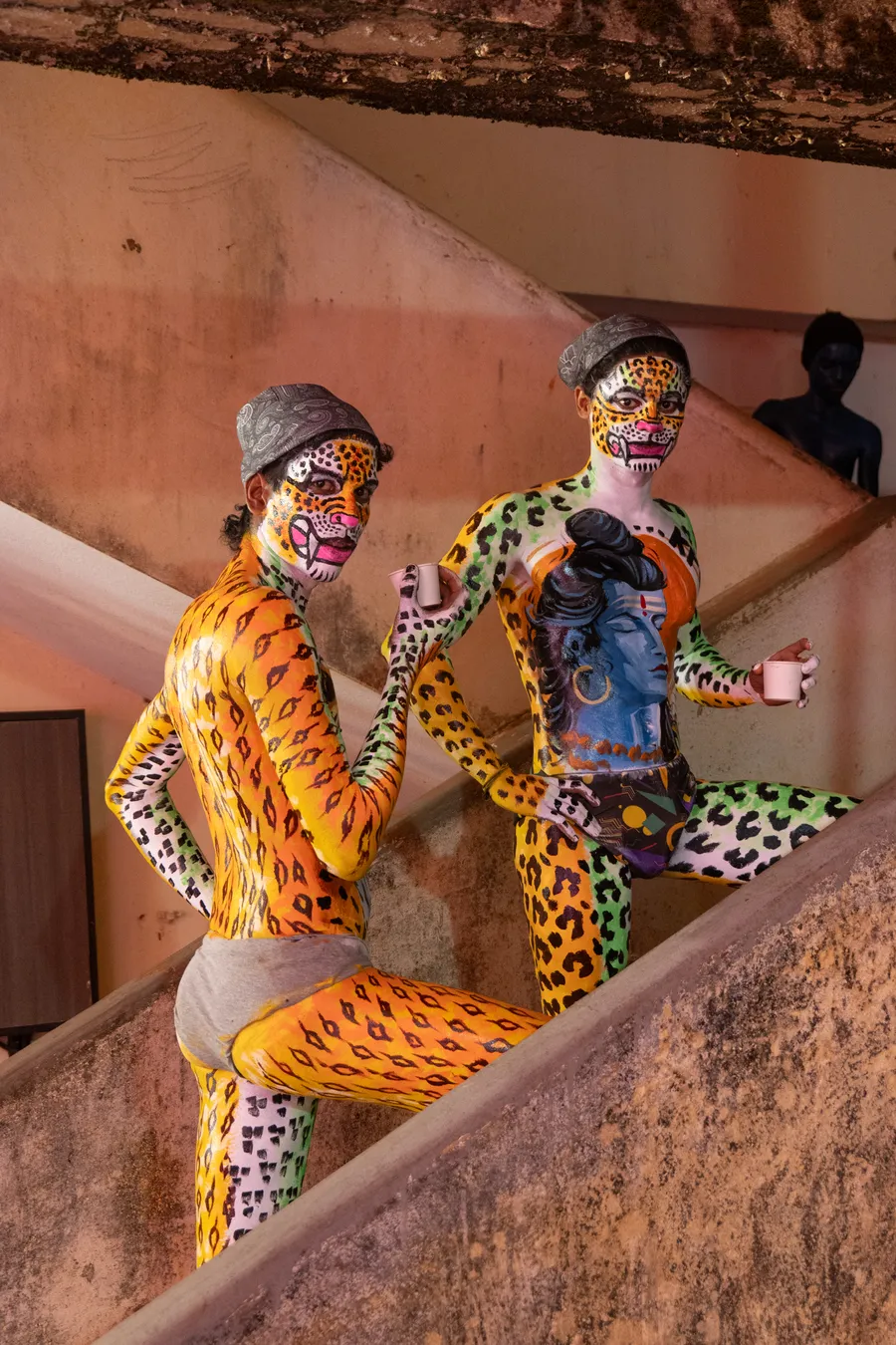 Two face-painted, spotted performers look toward the camera from a concrete stairway while holding paper cups.
