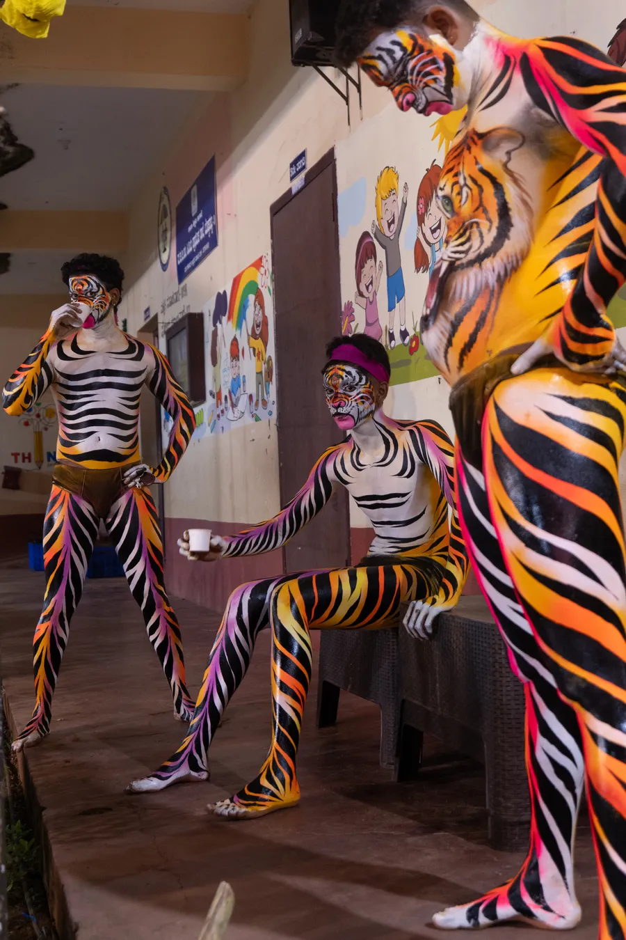 Three tiger-striped Pili Vesha performers pause with paper cups outside a building.