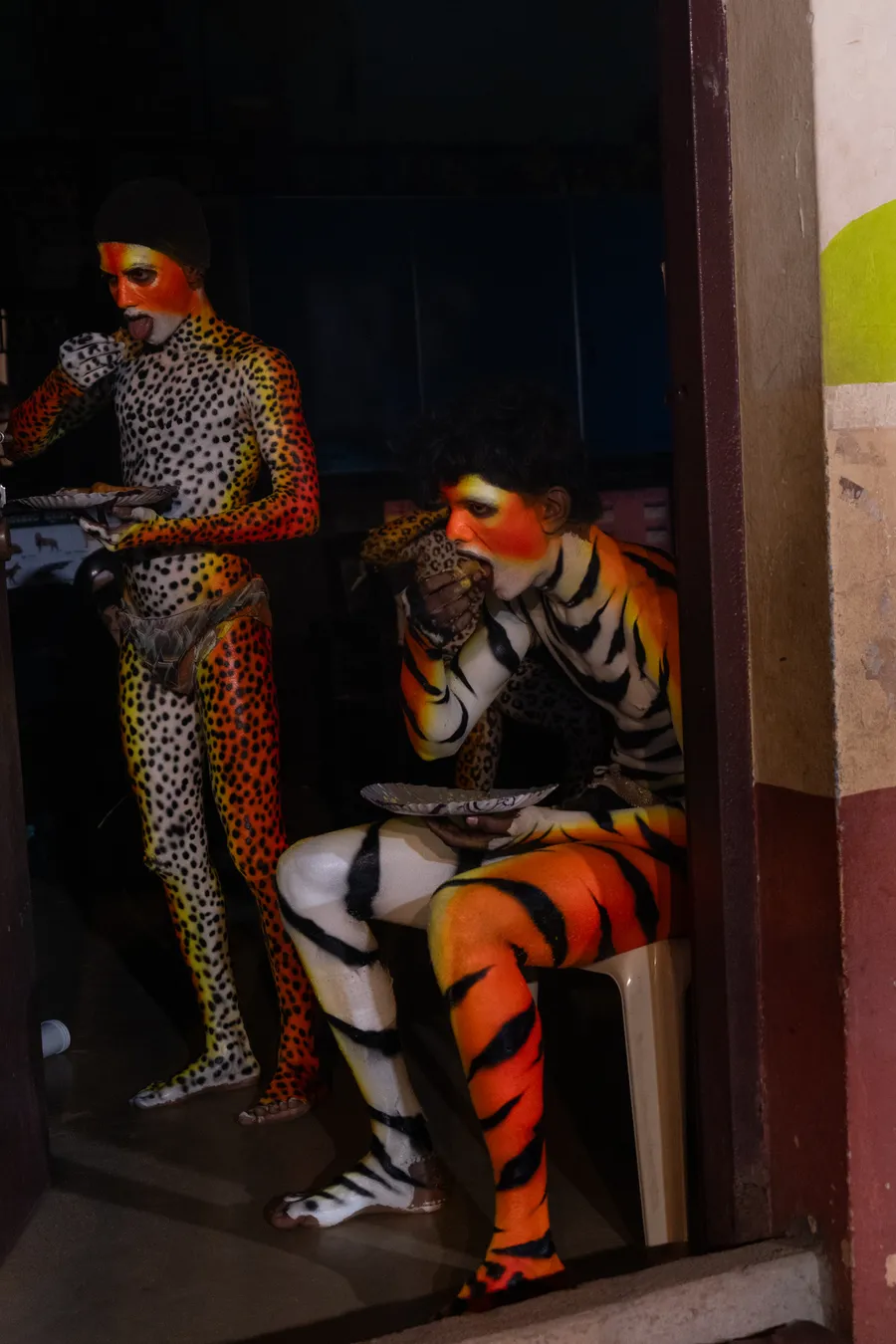 Two spotted and tiger-striped performers eat from paper plates in a doorway.