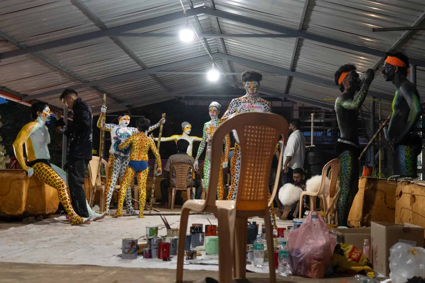 Body-painted performers and artists gather beneath a roof amid chairs, paint cans, and headpieces.