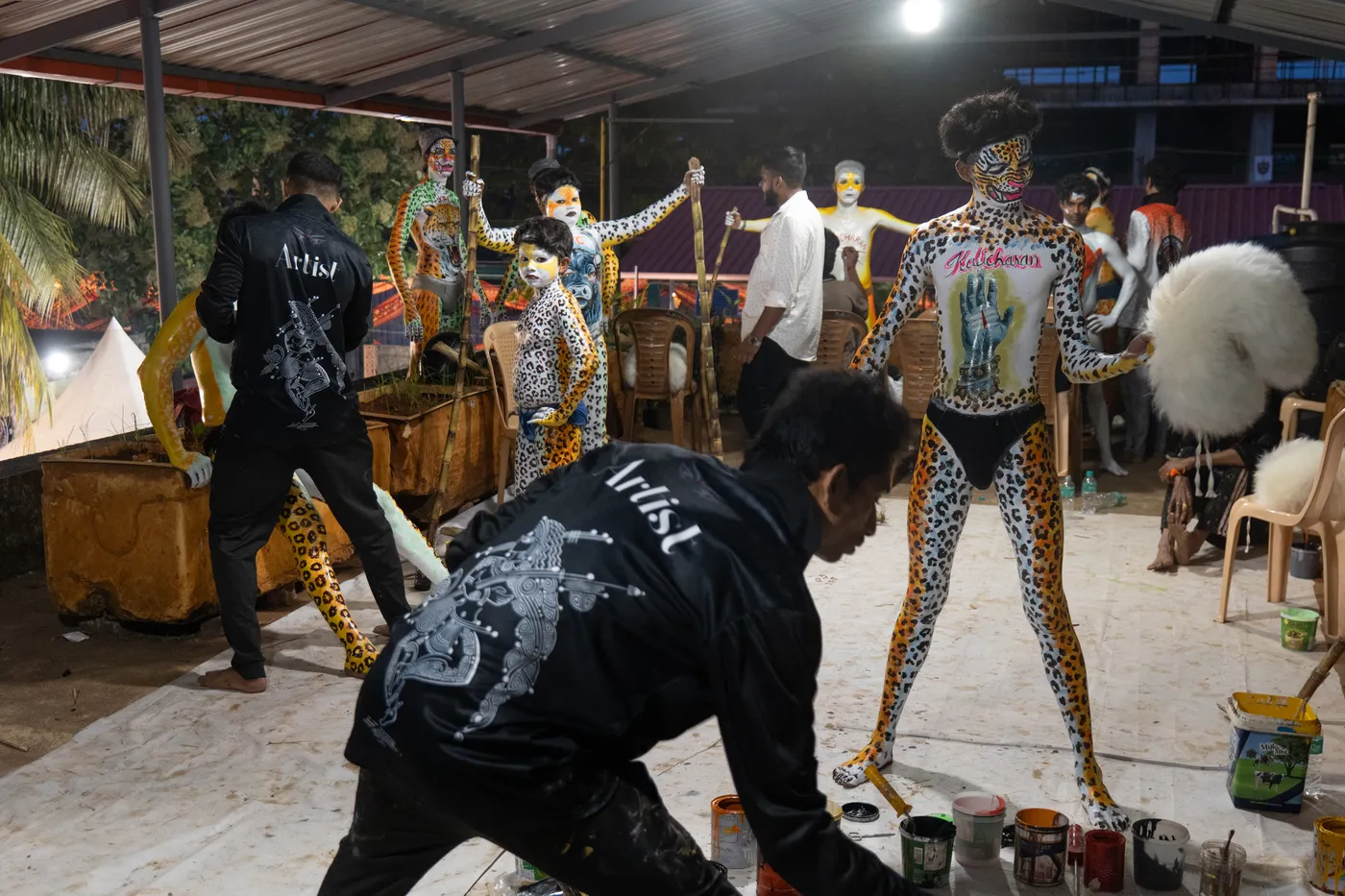 Artist crouches among paint cans while spotted and striped performers wait beneath a roof.