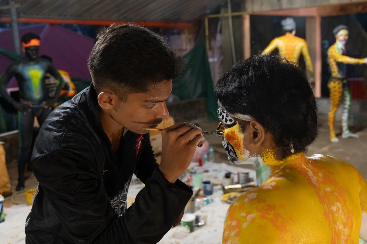 Close view of an artist painting black outlines on a performer’s yellow-and-white face design.