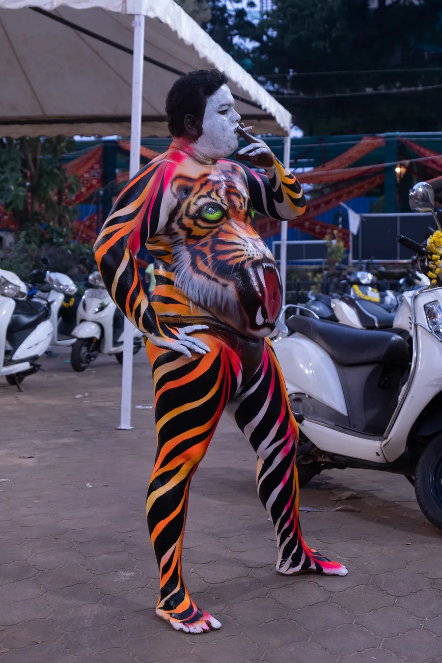 Tiger-striped performer with a painted tiger face on the torso smokes beside parked scooters.