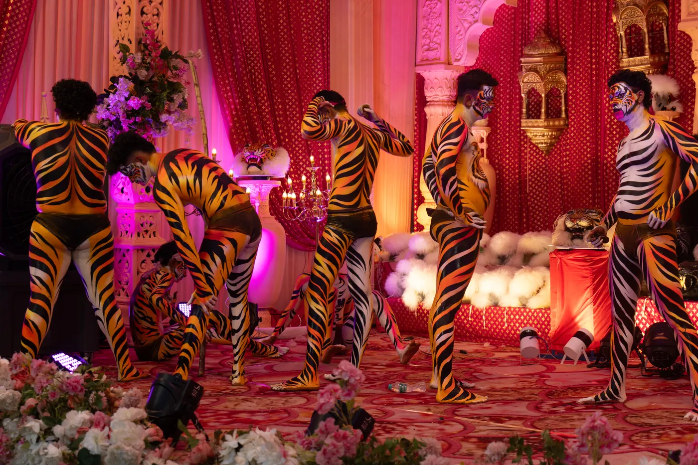 Several tiger-striped performers adjust body paint and costumes on a flower-decorated stage.