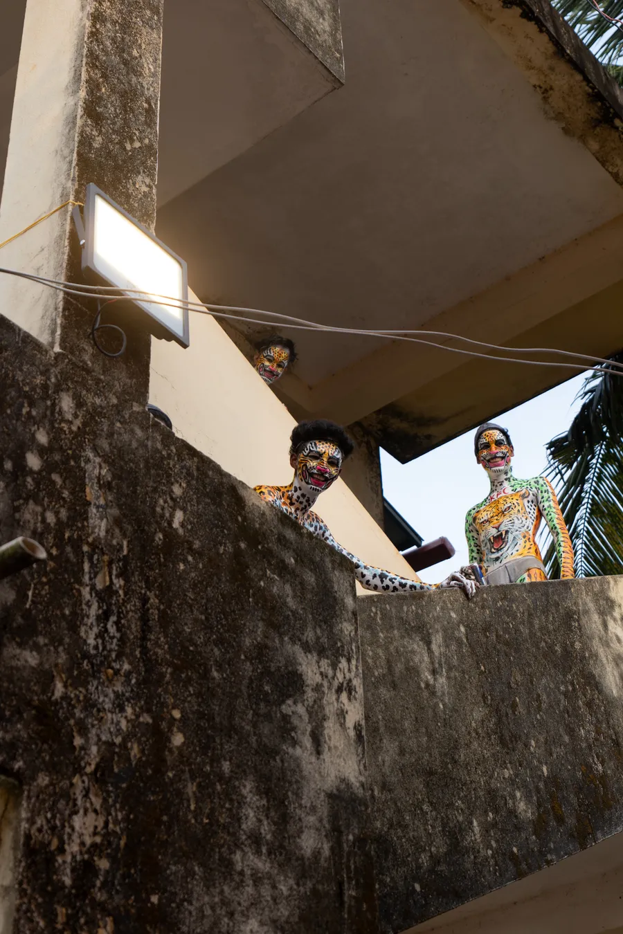 Three face-painted performers peer over a weathered concrete balcony.