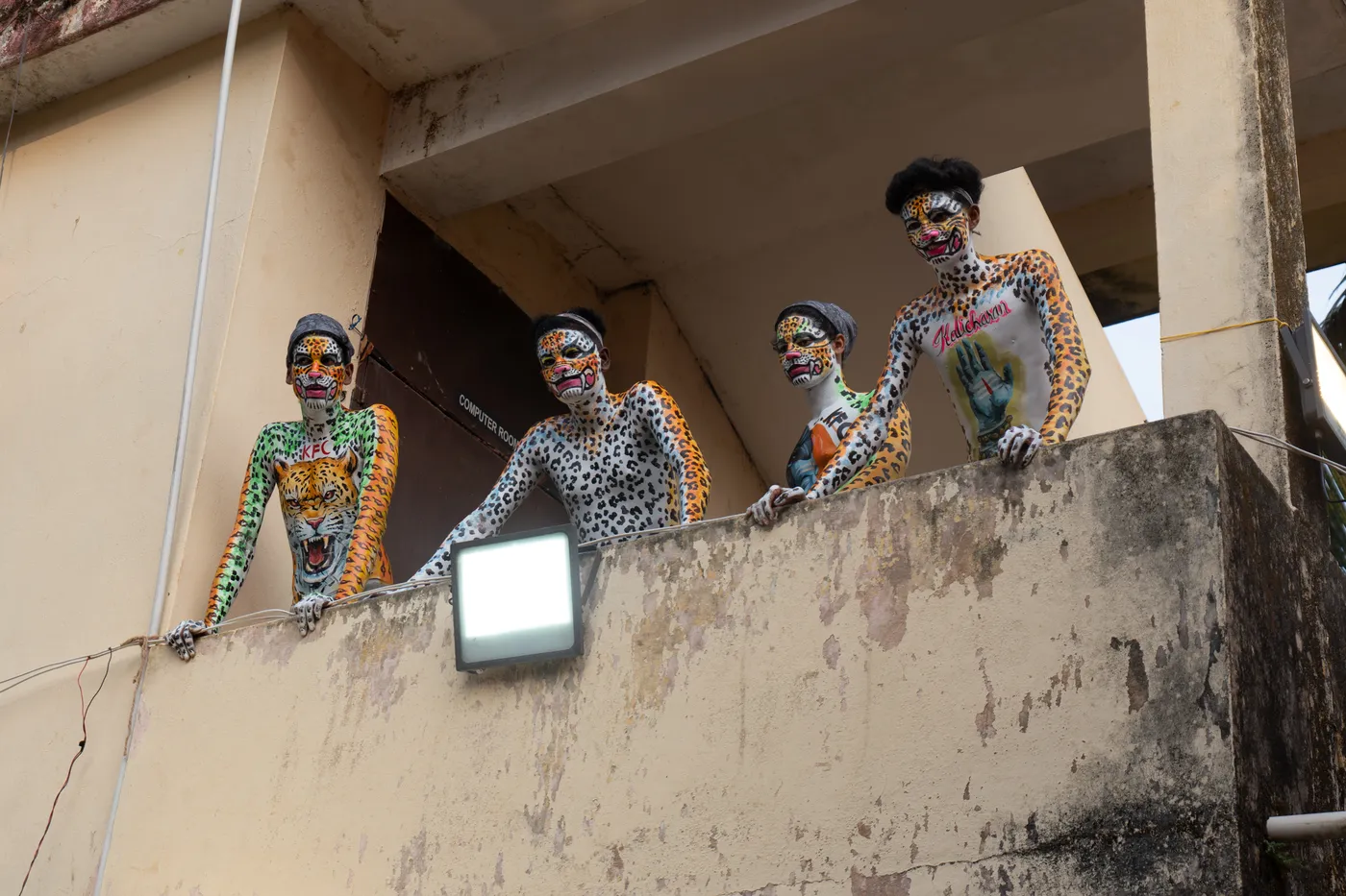 Four spotted body-painted performers lean over a concrete balcony.